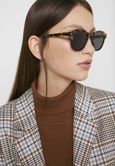 schildpaddenszonnebril met ronde glazen, dikke monturen en een glanzende afwerking, gecombineerd met een bruine geribbelde coltrui en een geblokte blazer.