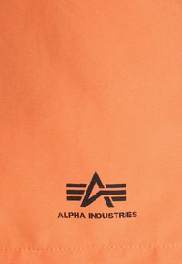 Orange stoff med en svart "Alpha Industries" logo og et geometrisk design. Glatt tekstur, minimalistisk stil og detaljert søm.