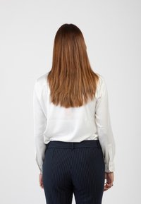 Camicia bianca a maniche lunghe con polsini abbottonati abbinata a pantaloni a righe blu navy. Capelli castano chiaro che ricadono oltre le spalle, visti da dietro.