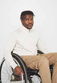 Bílý svetr na zip s ribbed texturou, béžové kalhoty, sedící na invalidním vozíku. Pozadí je jednoduché bílé, což zdůrazňuje subjekt.