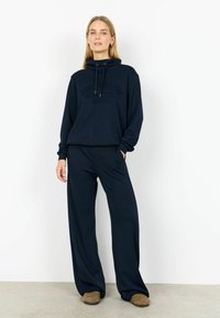 Costum sport bleumarin format dintr-un hanorac cu glugă, cu text pe partea din față, și pantaloni largi. Țesătura pare moale și texturată, oferind confort.