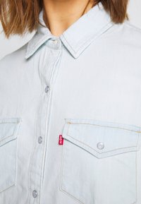 Light blue denim shirt with a pointed collar, button closure, two front pockets, and contrast stitching. Features a small red Levi's tag.