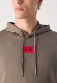 Felpa khaki con un patch rettangolare rosso che presenta la scritta "HUGO" in nero. Coulisse con punte metalliche e superficie in tessuto testurizzato.