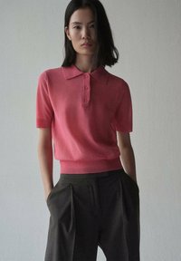 Polo shirt rosa a maniche corte con colletto classico e tre bottoni, abbinato a pantaloni scuri a pieghe per un look contemporaneo.