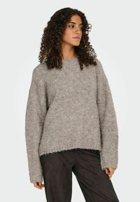 Femme aux cheveux bouclés portant un pull en tricot gris ample et un pantalon sombre, regardant sur le côté devant un fond uni.