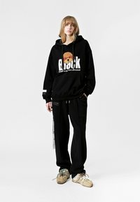 Sudadera negra con diseño gráfico y texto, combinada con pantalones deportivos negros. Zapatos en beige con cordones multicolores. Estilo minimalista, corte relajado.