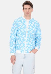 Jachetă bomber albastră deschis cu modele florale albe, având închidere cu fermoar și manșete și margine ribbat albă. Purtați cu pantaloni din denim deschis.