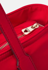 Czerwona torebka z gładką fakturą, z goldowym zamkiem błyskawicznym oraz logo "GUESS" na suwaku. Wnętrze ma jasne wykończenie.