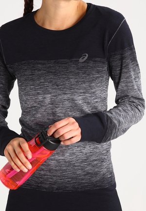 Femme portant un t-shirt de sport noir et gris à manches longues tenant une bouteille d'eau rose avec un bouchon noir.