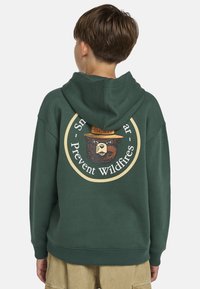 Grüne Kapuzenjacke aus Baumwolle mit einem runden Grafikdruck eines Bären, der einen Hut trägt, mit dem Text "Smokey" und dem Slogan "Waldbrände verhindern" auf der Rückseite.