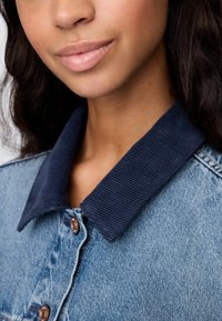 Jeans jakna s temno modrim ovratnikom iz korduroja, bronastim gumbnim zapiranjem in teksturirano modro tkanino z izpranim videzom.