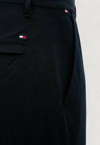 Primo piano di pantaloni neri con tasca laterale e piccolo logo rettangolare rosso, bianco e blu vicino alla tasca e alla vita.