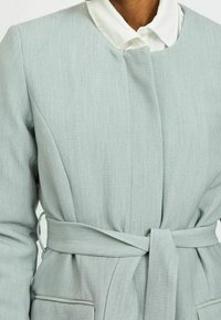 Cappotto grigio su misura con vita arricciata, scollo rotondo e maniche lunghe. Tessuto testurizzato con sottili dettagli di cucitura.