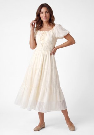 Vrouw poseert in een crèmekleurige midi-jurk met pofmouwen en ballerina’s tegen een effen witte achtergrond.