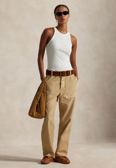 Woman wearing white sleeveless top, beige pants with large pockets, brown belt, brown shoes, black sunglasses, holding brown suede bag.