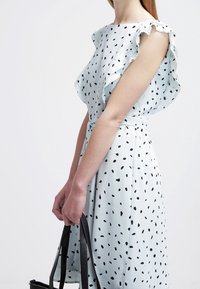 Femme portant une robe blanche avec des pois noirs irréguliers, des manches volantes, et tenant des anses de sac fourre-tout noir devant un fond blanc uni.