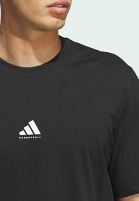 Camiseta de algodón negra con cuello redondo. Presenta un logo blanco de Adidas y el texto "BASKETBALL" en el pecho. Manga corta.