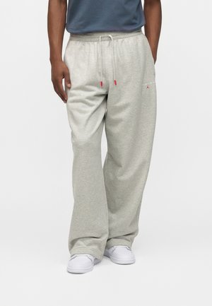 Mann trägt hellgraue lockere Jogginghose mit Kordelzug und weißen Sneakers, steht vor einem einfarbigen weißen Hintergrund.