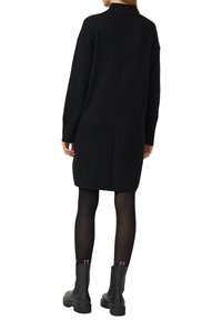 Schwarzes, grob gestricktes Oversize-Pulloverkleid mit hohem Kragen, langen Ärmeln und geripptem Muster. Kombiniert mit schwarzen Stiefeletten und Strumpfhosen.