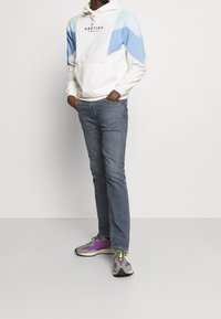 Vit hoodie med blå och ljusgrön geometrisk detalj och en framficka, kombinerad med blå jeans och flerfärgade sneakers.