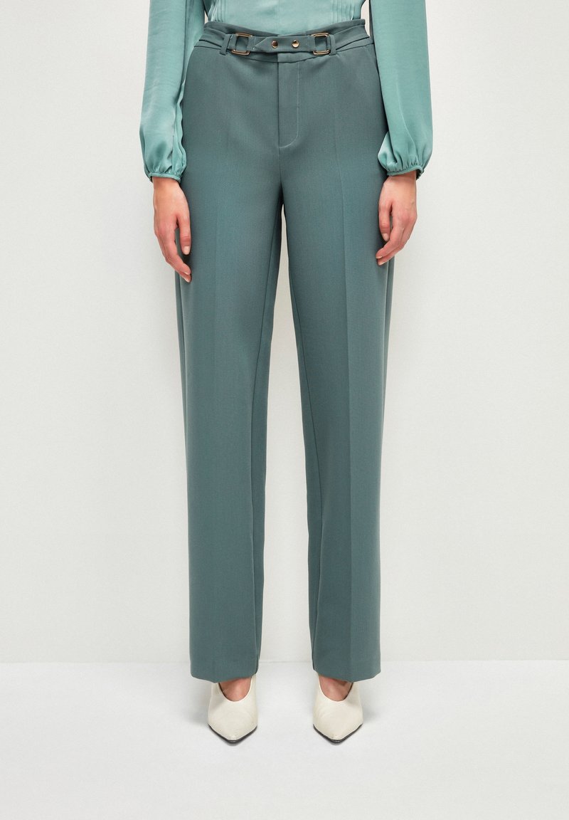 Pantalon slim bleu sarcelle avec ceinture assortie, porté avec un chemisier bleu sarcelle clair et des chaussures blanches à bout pointu sur un fond uni.