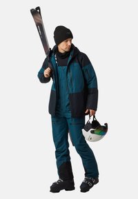 Man in een teal en zwarte skikleding houdt een helm met getinte vizier in één hand en heeft ski's over zijn schouder, draagt een zwarte beanie en skischoenen.