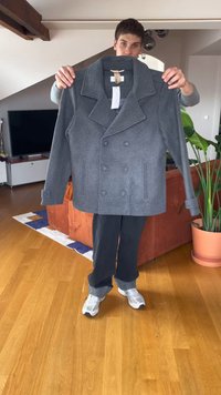 Person som håller en grå dubbelknäppt sjömansjacka med breda slag, stående i ett vardagsrum med trägolv och en krukväxt.
