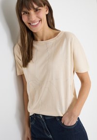 Femme souriante aux longs cheveux bruns, portant une chemise beige à manches courtes et un jean bleu foncé, debout avec une main dans la poche contre un mur blanc.