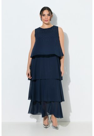 Vrouw draagt een mouwloze marineblauwe gelaagde maxi-jurk met versieringen op het lijfje, gecombineerd met zilveren hoge hakken.