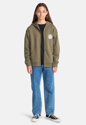 Adolescente che indossa un felpa con zip verde oliva, maglietta nera, jeans blu e sneakers in bianco e nero, in piedi contro uno sfondo neutro.