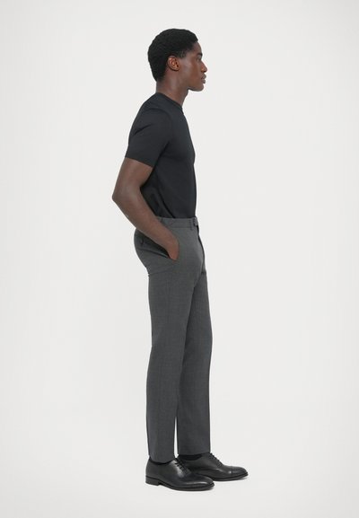 Camisa negra ajustada, pantalones grises de corte recto con bolsillos y zapatos de piel negros, mostrados de perfil sobre un fondo blanco.