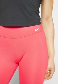 Różowe legginsy elastyczne z wysokim stanem, gładką teksturą i białym logo Nike z lewej strony. Czarny krótki top powyżej.