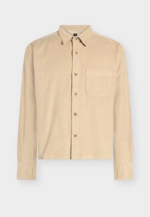 Hosszú ujjú bézs kordbársony ing gallérral, gombos elülső résszel és egyetlen zsebbel a bal mellrészen. Texturált anyag részlet.