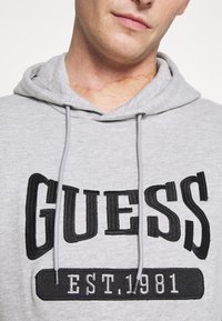 Sudadera gris con un gran logo negro bordado de "GUESS" y "EST. 1981" en el dobladillo, con cordón y tela texturizada.