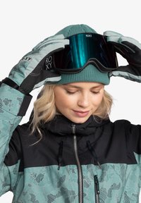 Türoser und schwarzer Schneejacke mit gemusterten Akzenten, grüner Strickmütze und dunklen Skibrillen mit reflektierenden Gläsern, gehalten von Handschuhen.