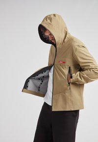 Chaqueta marrón claro con capucha, que presenta un logo rojo, bolsillos con cremallera y un forro negro. El tejido parece suave y resistente al agua.
