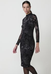 Rochie neagră cu imprimeu floral, cu guler înalt, mâneci lungi dintr-un material transparent și o siluetă ajustată. Material moale, asemănător catifelei, cu accente texturate.