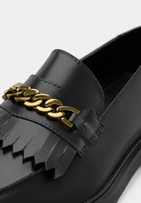 Mocassin en cuir noir avec un design à franges et un accent de chaîne dorée sur le devant. Texture lisse avec des coutures détaillées.