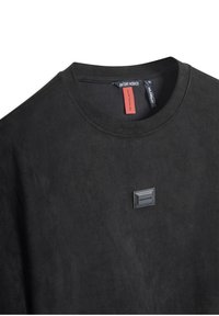 Crni suede dukserica s okruglim izrezom i glatkom teksturom. Ima mali pravokutni logotip na prednjoj strani i oznaku s brendom na vratu.