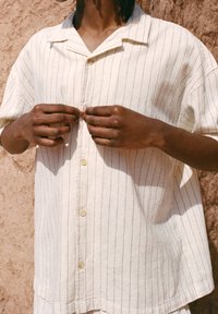 Chemise beige clair à manches courtes avec des rayures verticales, dotée d'un col cranté et d'une rangée de boutons. Le tissu semble texturé.