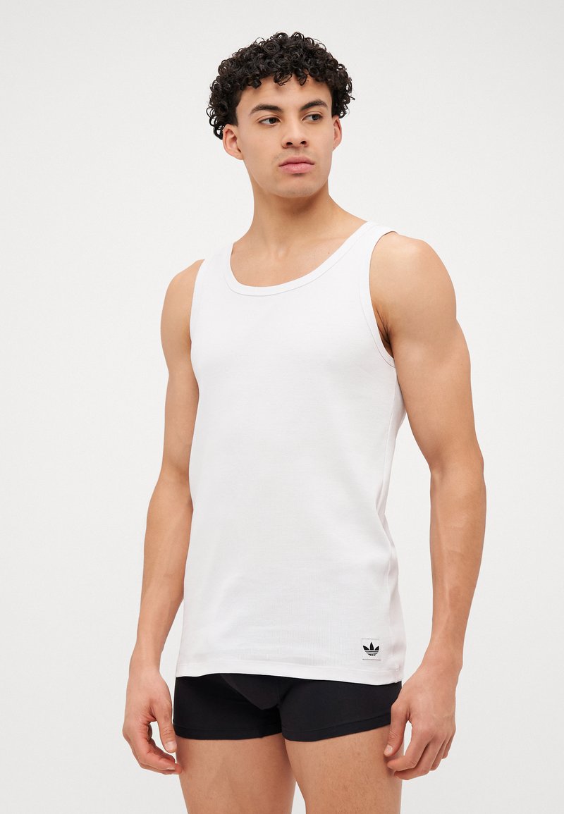 Λευκό βαμβακερό σορτς με ριβέ σχέδιο, στρογγυλή λαιμόκοψη και ένα μικρό μαύρο λογότυπο στο κάτω αριστερό μέρος. Μοντέλο που φοράει μαύρα σόρτς.