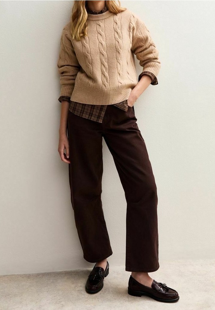 Femme portant un pull torsadé beige sur une chemise à carreaux marron, un pantalon large marron foncé et des mocassins marron foncé, debout contre un mur blanc.