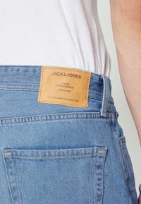 Jeans de denim azul claro com uma etiqueta de couro na parte de trás da cintura, com o nome da marca e texto em relevo. Design clássico com cinco bolsos.