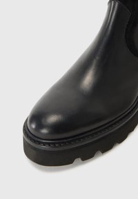 Bottine en cuir noir avec un bout arrondi, présentant un panneau en daim texturé et une semelle en caoutchouc épaisse et nervurée. Détails de couture visibles.