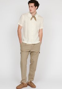 Camisa de lino beige de manga corta con cuello, combinada con pantalones beige claros y zapatos de ante marrones. Presenta una bufanda de diseño en el cuello.