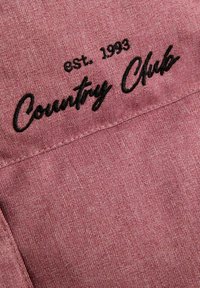 Zbliżenie różowego materiału z czarnym haftowanym napisem "est. 1993 Country Club" na szwie.