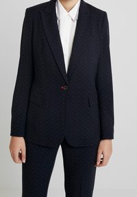 Blazer bleu marine avec un motif discret de petits points, fermeture à un bouton, deux poches frontales, coupe ajustée et revers nets. Porté sur une chemise blanche.