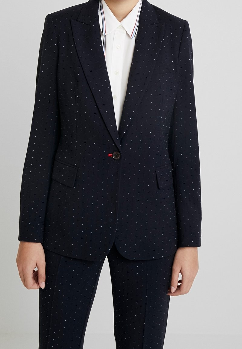 Blazer bleu marine avec un motif discret de petits points, fermeture à un bouton, deux poches frontales, coupe ajustée et revers nets. Porté sur une chemise blanche.