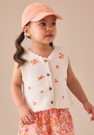 Young girl wearing a peach cap, white sleeveless cardigan with pink floral embroidery, and a pink floral skirt, looking slightly to the side.