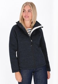 Jauna moteris, vilkinti tamsiai mėlyną zip-up flisinę striukę su balta šeršelu, mėlynas džinsus ir dryžuotą marškinėlį, žiūrinti į dešinę su nedideliu šypsniu.
